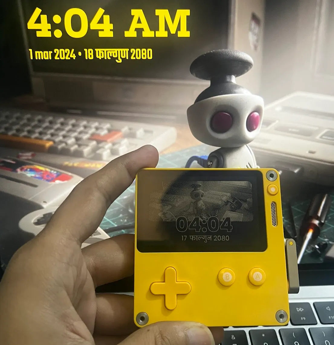 The Playdate in hand showing 04:04 and 17 फाल्गुण 2080, in front of the same robot wallpaper on a monitor